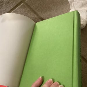 Puede incluir: Un libro de tapa dura con una cubierta verde brillante est&aacute; abierto, revelando p&aacute;ginas blancas. El lomo del libro es visible, y una mano est&aacute; parcialmente a la vista, sosteniendo el libro abierto. El libro est&aacute; sobre una superficie embaldosada.