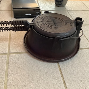 May include: A vintage cast iron waffle iron with a circular cooking surface and long, spring-handled grips. The iron is dark brown and shows signs of age and use. The waffle iron is designed to be used over a stovetop or open fire.