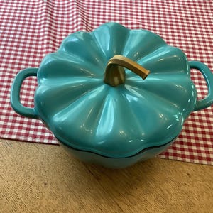 May include: A teal pumpkin-shaped casserole dish with a gold-coloured stem and handles. The dish sits on a red and white checkered tablecloth and a wooden surface.