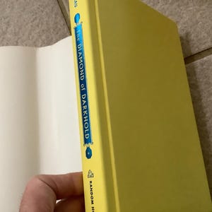 Puede incluir: Un libro de tapa dura titulado "The Diamond of Darkhold" con una cubierta y lomo de color amarillo brillante. El lomo presenta el t&iacute;tulo en letras azules y blancas, con el logotipo de Random House en la parte inferior. El libro est&aacute; parcialmente abierto, revelando p&aacute;ginas blancas.