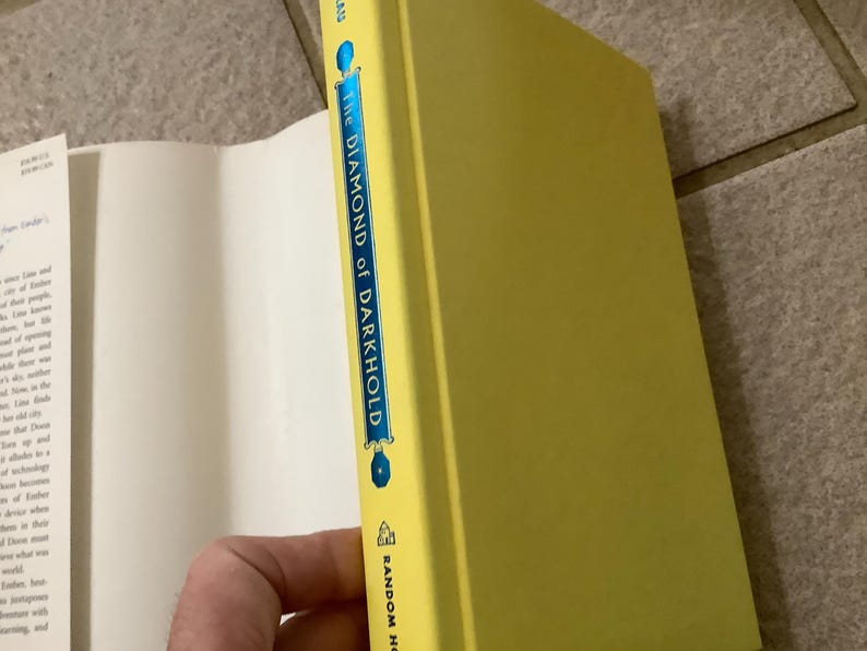 Puede incluir: Un libro de tapa dura titulado "The Diamond of Darkhold" con una cubierta y lomo de color amarillo brillante. El lomo presenta el t&iacute;tulo en letras azules y blancas, con el logotipo de Random House en la parte inferior. El libro est&aacute; parcialmente abierto, revelando p&aacute;ginas blancas.