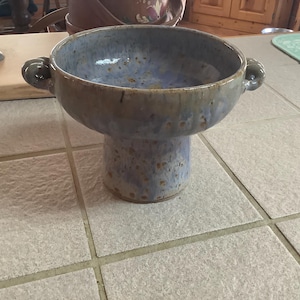May include: A handcrafted ceramic bowl with a pedestal base. The bowl is a muted blue with brown accents and two small handles. The bowl is approximately 8 inches in diameter and 6 inches tall. Perfect for serving soups or salads.