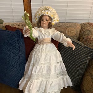 May include: A porcelain doll in a white dress with lace trim and a floral headpiece. The doll holds a sprig of greenery and has a tan sash. The doll's dress has multiple layers and ruffles. The doll is wearing white shoes.