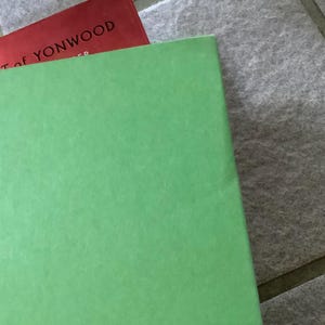 Puede incluir: Primer plano de dos libros. El libro superior es rojo, con el t&iacute;tulo "ART OF YONWOOD" visible. El libro inferior es de color verde claro liso. Los libros descansan sobre una superficie de baldosas grises.