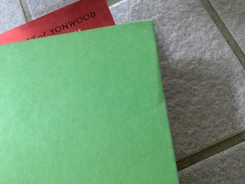 Puede incluir: Primer plano de dos libros. El libro superior es rojo, con el t&iacute;tulo "ART OF YONWOOD" visible. El libro inferior es de color verde claro liso. Los libros descansan sobre una superficie de baldosas grises.