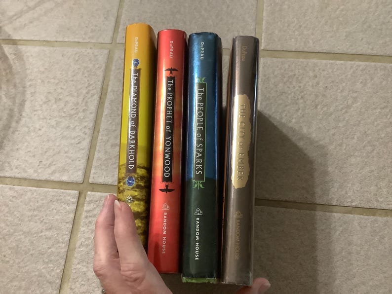 Puede incluir: Cuatro libros de tapa dura con lomos coloridos son sostenidos por una mano. Los libros se titulan "The Diamond of Darkhold", "The Prophet of Yonwood", "The People of Sparks" y "The City of Ember". Los libros son publicados por Random House.
