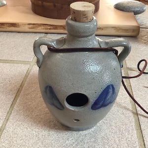 May include: A gray ceramic jug with a cork stopper, two handles, and a dark brown leather strap. The jug features two blue leaf-like designs and a large circular opening. A small hole is visible near the bottom.