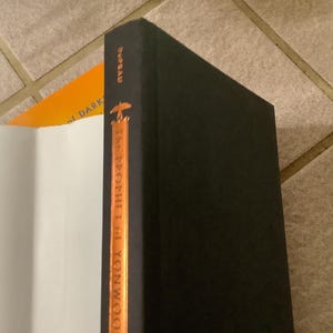 Puede incluir: Una pila de libros, con un libro de tapa dura negro en primer plano. La columna vertebral del libro negro tiene el t&iacute;tulo "The Prophet of Yonwoo" en letras doradas. Un libro abierto con p&aacute;ginas blancas y un libro naranja son visibles.