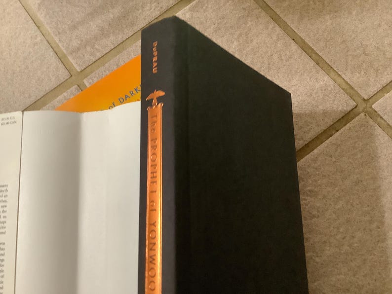 Puede incluir: Una pila de libros, con un libro de tapa dura negro en primer plano. La columna vertebral del libro negro tiene el t&iacute;tulo "The Prophet of Yonwoo" en letras doradas. Un libro abierto con p&aacute;ginas blancas y un libro naranja son visibles.