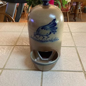 May include: A tan ceramic water dispenser with a blue bird design. The dispenser has a dark blue top and a small bowl at the bottom. The background includes a table and a vase of flowers.