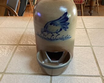 May include: A tan ceramic water dispenser with a blue bird design. The dispenser has a dark blue top and a small bowl at the bottom. The background includes a table and a vase of flowers.