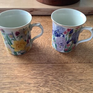 May include: Two ceramic mugs with floral designs. One mug features yellow daffodils and other flowers, while the other showcases purple irises and red flowers. Both mugs have gold trim and white handles, set on a wooden surface.