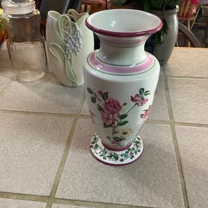 Może przedstawiać: Biały ceramiczny wazon z rozszerzoną krawędzią i podstawą. Wazon ma różowy pasek wokół szyi i podstawy, z kwiatowym wzorem różowych róż i zielonych liści. Wazon ma około 25 cm wysokości.