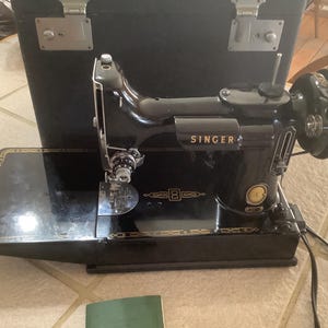 Può includere: Macchina da cucire Singer nera con dettagli dorati, presentata con una custodia per il trasporto. La macchina ha una finitura lucida ed è posizionata su una superficie piastrellata. La parola "SINGER" è visibile sulla macchina.