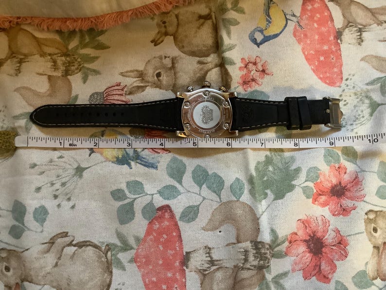 May include: A silver-toned wristwatch with a black leather band. The watch face has a white background with black markings and a silver-toned logo. The watch is 5 inches long.