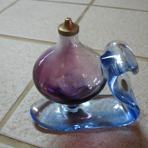 May include: A purple glass bottle with a gold top sits on a blue glass base. The bottle is shaped like a bulb and the base is shaped like a wave.