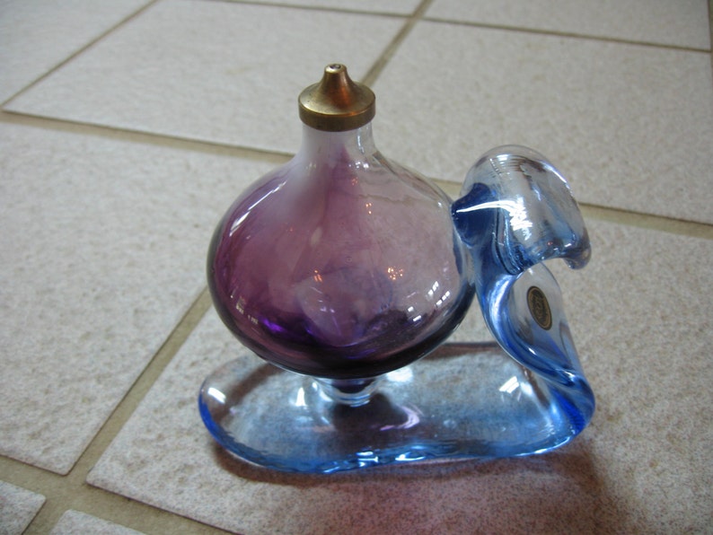 May include: A purple glass bottle with a gold top sits on a blue glass base. The bottle is shaped like a bulb and the base is shaped like a wave.