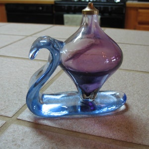 May include: A purple glass bottle with a blue glass base. The bottle is shaped like a teardrop and has a small opening at the top. The base is shaped like a swan.