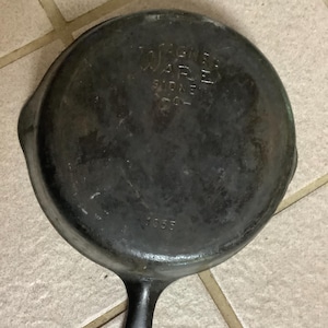 May include: A round black cast iron skillet with a long handle. The underside features embossed text reading Wagner Ware Sidney -O- and the number 1055. The surface shows signs of wear and discoloration consistent with a seasoned metal cooking pan.