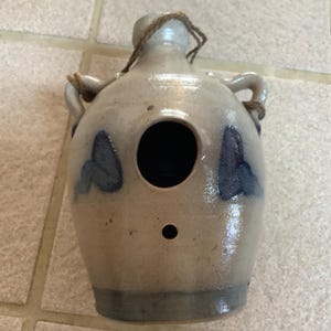 Op de afbeelding: Een keramisch vogelhuisje in de vorm van een kruik met een blauw en wit vlinderontwerp. Het vogelhuisje heeft een ronde opening waar vogels doorheen kunnen.