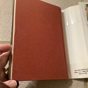 Puede incluir: Un libro abierto con una cubierta roja y p&aacute;ginas blancas. El lomo tiene un borde dorado. El libro se sostiene abierto, revelando texto y una foto de una mujer en la p&aacute;gina derecha. El texto "Made in USA" es visible.