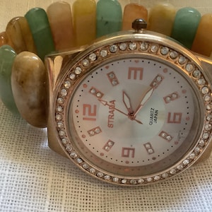 Könnte beinhalten: Eine goldfarbene Armbanduhr mit weißem Zifferblatt und einer mit Kristallen besetzten Lünette. Die Uhr ist an einem mehrfarbigen Stein-Armband befestigt. Das Zifferblatt der Uhr zeigt "Straia Quartz Japan".