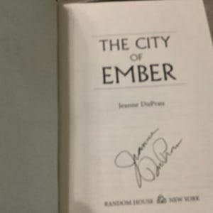 Puede incluir: Libro abierto con el t&iacute;tulo "THE CITY OF EMBER" en fuente negra. El nombre del autor, Jeanne DuPrau, est&aacute; debajo. Hay una firma presente. El logotipo de Random House est&aacute; en la parte inferior.