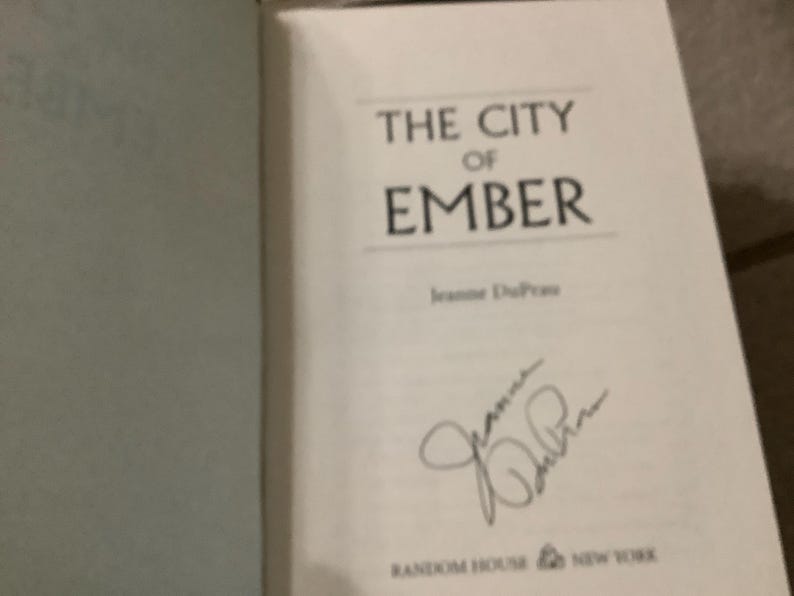Puede incluir: Libro abierto con el t&iacute;tulo "THE CITY OF EMBER" en fuente negra. El nombre del autor, Jeanne DuPrau, est&aacute; debajo. Hay una firma presente. El logotipo de Random House est&aacute; en la parte inferior.