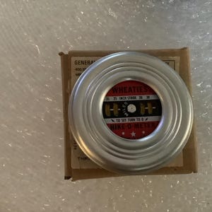 May include: A vintage Hike-O-Meter, a circular device with a silver rim, sits atop a cardboard box. The meter's face features the word "WHEATIES" and measurements for 63.5 cm strides. The text "HIKE-O-METER" is also visible.