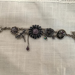 May include: Silver-toned bracelet featuring various charms, including a dragonfly, flowers, and a circular charm with a purple gem. The bracelet has a toggle clasp and a mix of dark blue and purple accents.