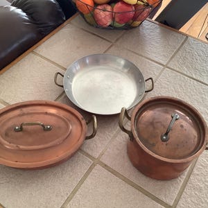 Puede incluir: Tres ollas de cocina, dos de cobre y una de plata, con asas. Las ollas de cobre tienen tapas.