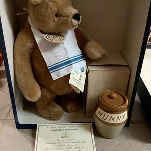 May include: A Winnie-the-Pooh plush bear with a bib and a honey pot. The bear is in a box with a certificate of registration. The honey pot is brown and beige with the word "HUNNY" on it.