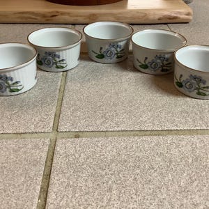 Puede incluir: Conjunto de cinco ramequines de cerámica blanca con un diseño floral. Cada ramequín presenta un patrón floral azul y verde con un borde dorado.