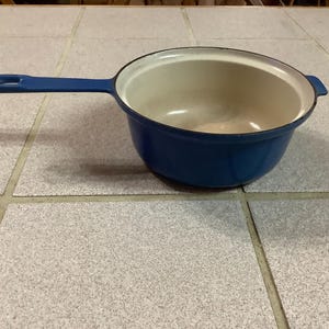 Puede incluir: Una cacerola azul de hierro fundido esmaltado con un mango largo y un interior blanco. La cacerola tiene forma redondeada y un pequeño asa en el lado opuesto al mango largo. La cacerola está sobre una superficie embaldosada.