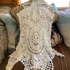 May include: White crocheted lace doily with a scalloped edge and a delicate floral pattern. The doily is laid out on a brown couch.