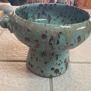 May include: A turquoise ceramic bowl with a pedestal base and two decorative handles. The bowl has a speckled pattern of brown and turquoise, and is likely handmade. This decorative bowl is suitable for serving or display.
