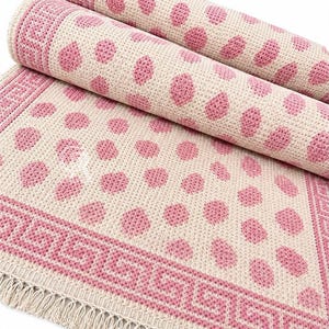 May include: A woven rug featuring a cream-colored base with a repeating pattern of pink polka dots. The rug has a wide border with a pink Greek key design and a fringe edge. The rug is partially rolled, showing the texture of the woven fabric.