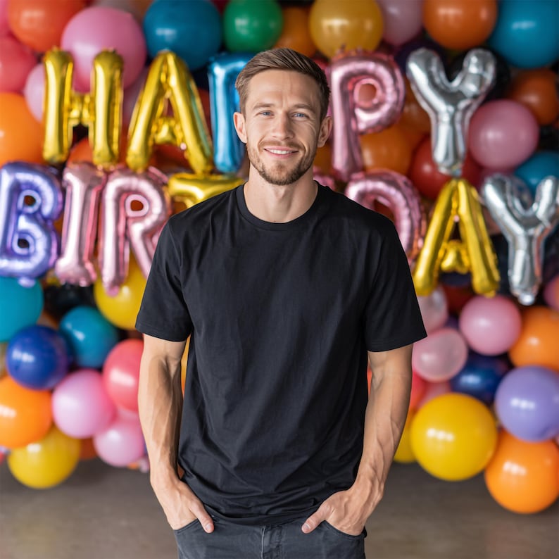 May include: A person wearing a black t-shirt stands in front of a colorful balloon backdrop. The balloons spell out "HAPPY BIRTHDAY" in gold, silver, and pink. The person is smiling, with hands in pockets.