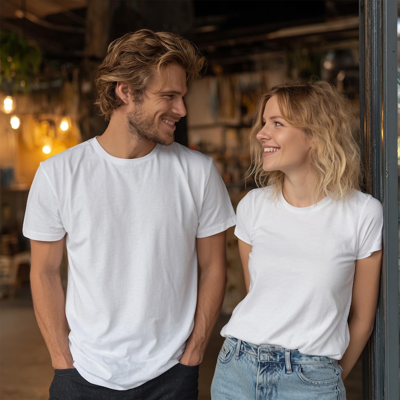 May include: Two people wearing white crew neck t-shirts and blue jeans. The man has his hands in his pockets and is smiling. The woman is leaning against a door frame and smiling. The background is blurred with warm lighting.
