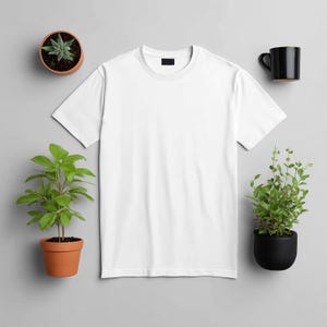 May include: A plain white t-shirt with a black tag on the collar is the focal point of this image. The shirt is surrounded by small potted plants and a black coffee mug, all set against a gray background. The plants are in terracotta and black pots.