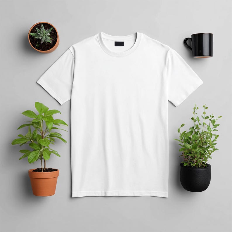 May include: A plain white t-shirt with a black tag on the collar is the focal point of this image. The shirt is surrounded by small potted plants and a black coffee mug, all set against a gray background. The plants are in terracotta and black pots.