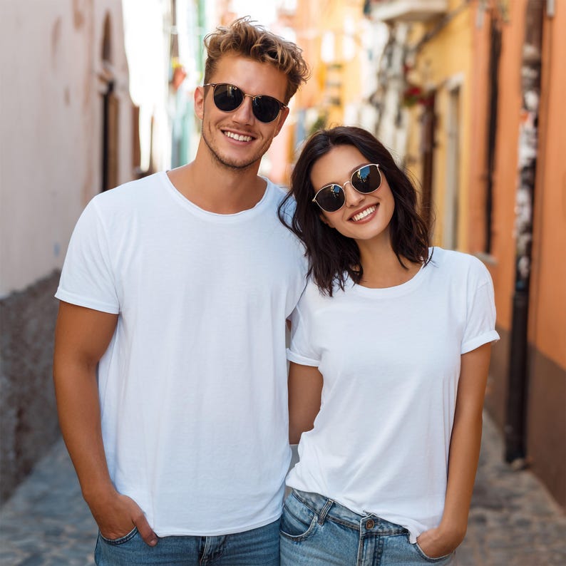 May include: A man and a woman wearing white t-shirts and sunglasses. The man has blond hair and the woman has dark hair. They are smiling and standing close together in a narrow street with colorful buildings.
