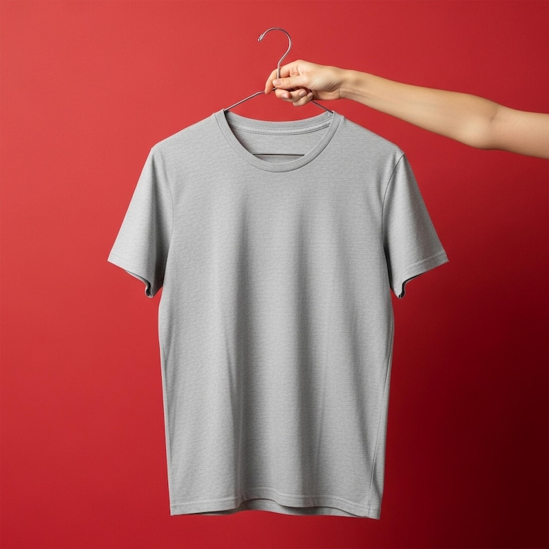 May include: A light gray, short-sleeved t-shirt hanging from a metal hanger. The shirt has a crew neck and is made of a soft, textured fabric. The background is a solid red color.