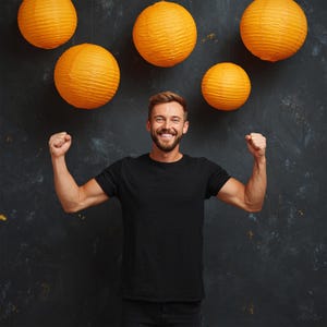May include: A man in a black t-shirt smiles with his fists clenched, with five orange paper lanterns hanging above him against a dark background. The lanterns are round and textured.