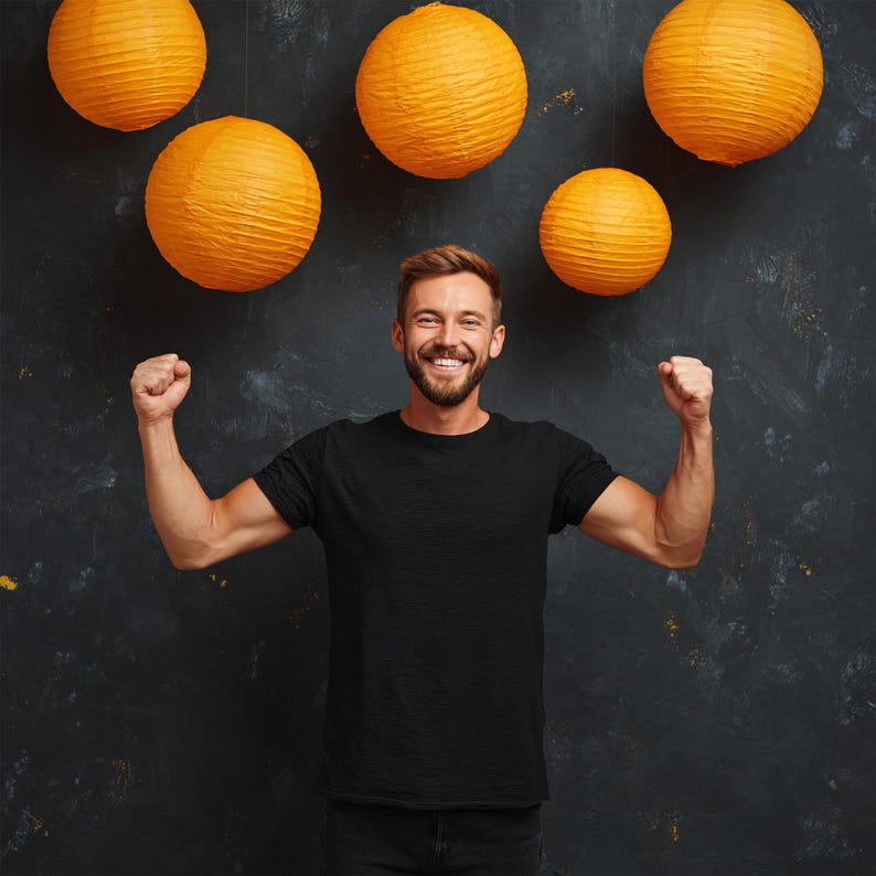 May include: A man in a black t-shirt smiles with his fists clenched, with five orange paper lanterns hanging above him against a dark background. The lanterns are round and textured.