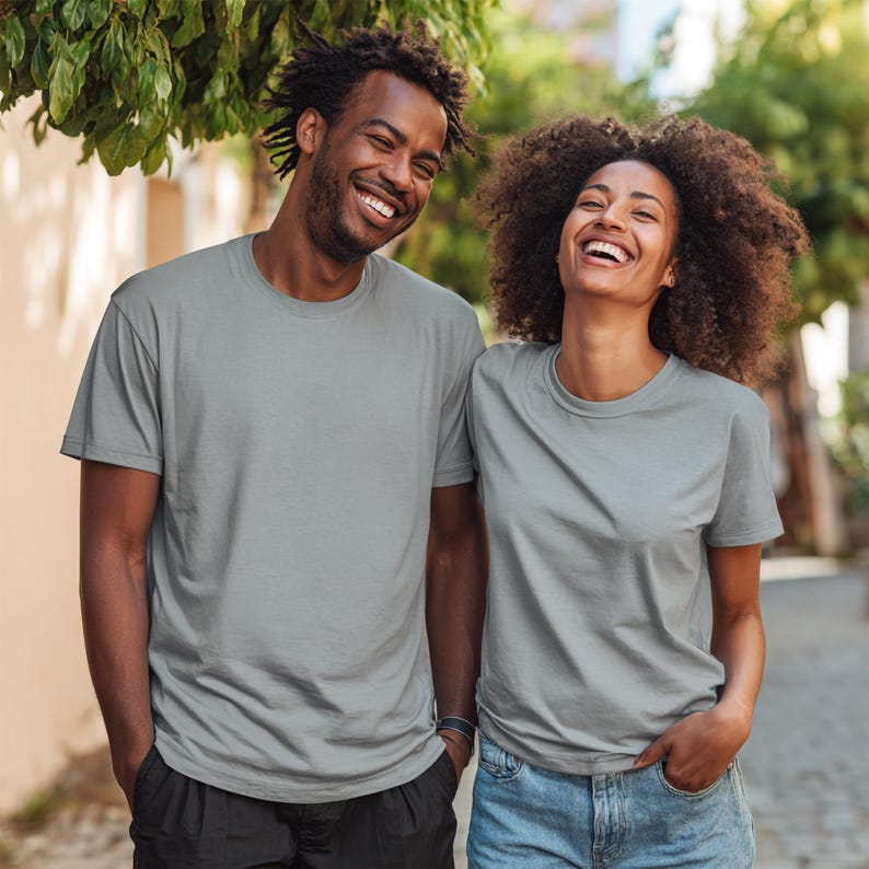 May include: Two people wearing gray crew neck t-shirts. The t-shirts are a solid, light gray color. The people are smiling and standing outside. The background is blurred, with green foliage and a light-colored wall visible.