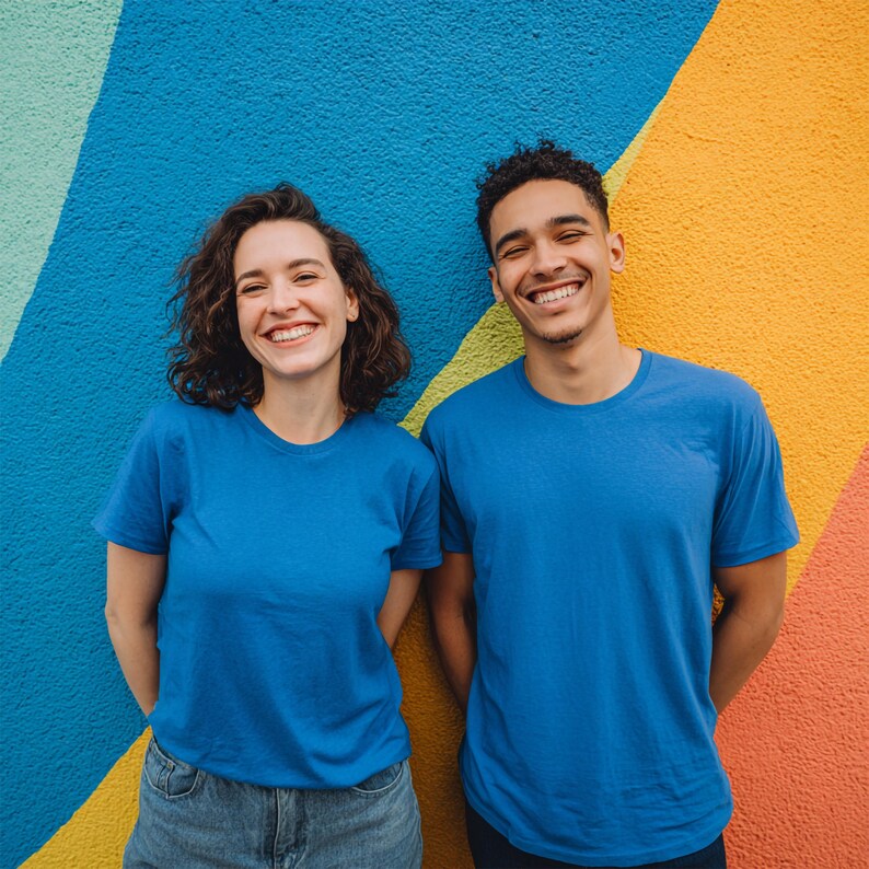 May include: Two people wearing blue crew neck t-shirts and denim jeans smile in front of a colorful wall. The wall has a vibrant design with blue, yellow, orange, and red sections.