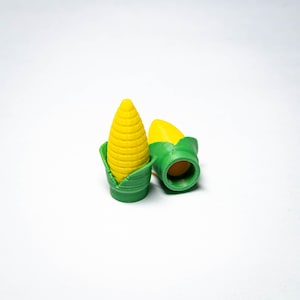 May include: Two small, three-dimensional corn-shaped caps featuring a bright yellow, textured kernel top and a green, leaf-like base. One cap stands upright, while the other lies on its side, revealing a hollow, cylindrical opening at the bottom.