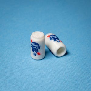 Könnte beinhalten: Zwei weiße, zylindrische Perlen mit blauen und roten Cartoon-Grafiken. Eine Perle steht aufrecht, die andere liegt auf der Seite. Der Hintergrund ist einfarbig hellblau.