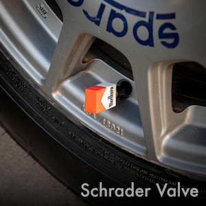 May include: A Schrader valve with a custom Marlboro design is installed on a silver car wheel. The valve cap features a red and white color scheme with the Marlboro logo. The tire is black, and the wheel has blue lettering.